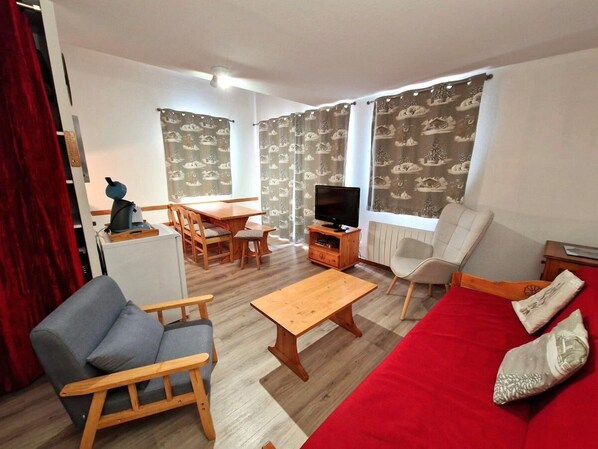 Ein Wohnzimmer mit einem roten Sofa, einem grauen Sessel, einem Holztisch, einem Fernseher und einem Essbereich mit Tisch und Stühlen.