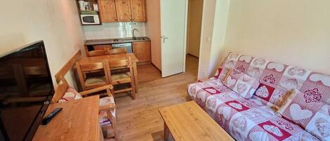 A compact living space with a wooden dining table, a sofa with patterned cushions, a television, and a kitchenette with wooden cabinets and a microwave.