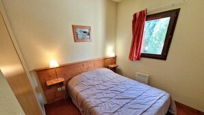 Quarto com cama, mesa de cabeceira, luminária na parede, janela com cortinas e radiador.