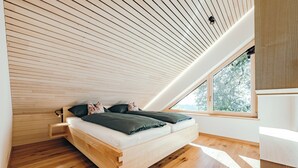 Un dormitori tipus loft modern amb sostre inclinat, un llit de fusta i un gran finestral que deixa entrar molta llum natural.