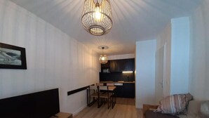 A compact living space with a flat-screen TV, a sofa, a dining table with chairs, a kitchen area, and a hanging light fixture.