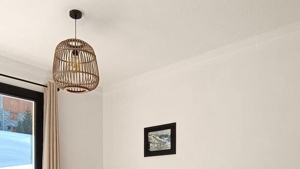 A modern living room with a wooden floor, a large window with curtains, a wooden chair, a TV on a wooden stand, and a hanging light fixture.