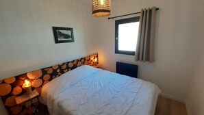 A bedroom with a bed, a headboard with a log pattern, a bedside table with a candle, a window with curtains, and a wall-mounted picture.