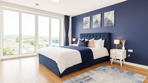 A modern bedroom with a large bed, a nightstand, a lamp, and a view of the city through large windows. - Luxury Penthouse in Harrow | 2BR | Gym | Games | Lounge | Free Parking (Harrow)