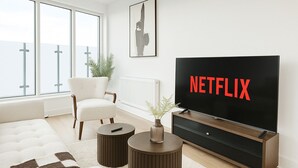 A modern living room with a large flat-screen TV displaying "NETFLIX," a white armchair, a round coffee table, and a view of the outdoors through large windows. - Luxury Penthouse in Harrow | 2BR | Gym | Games | Lounge | Free Parking (Harrow)