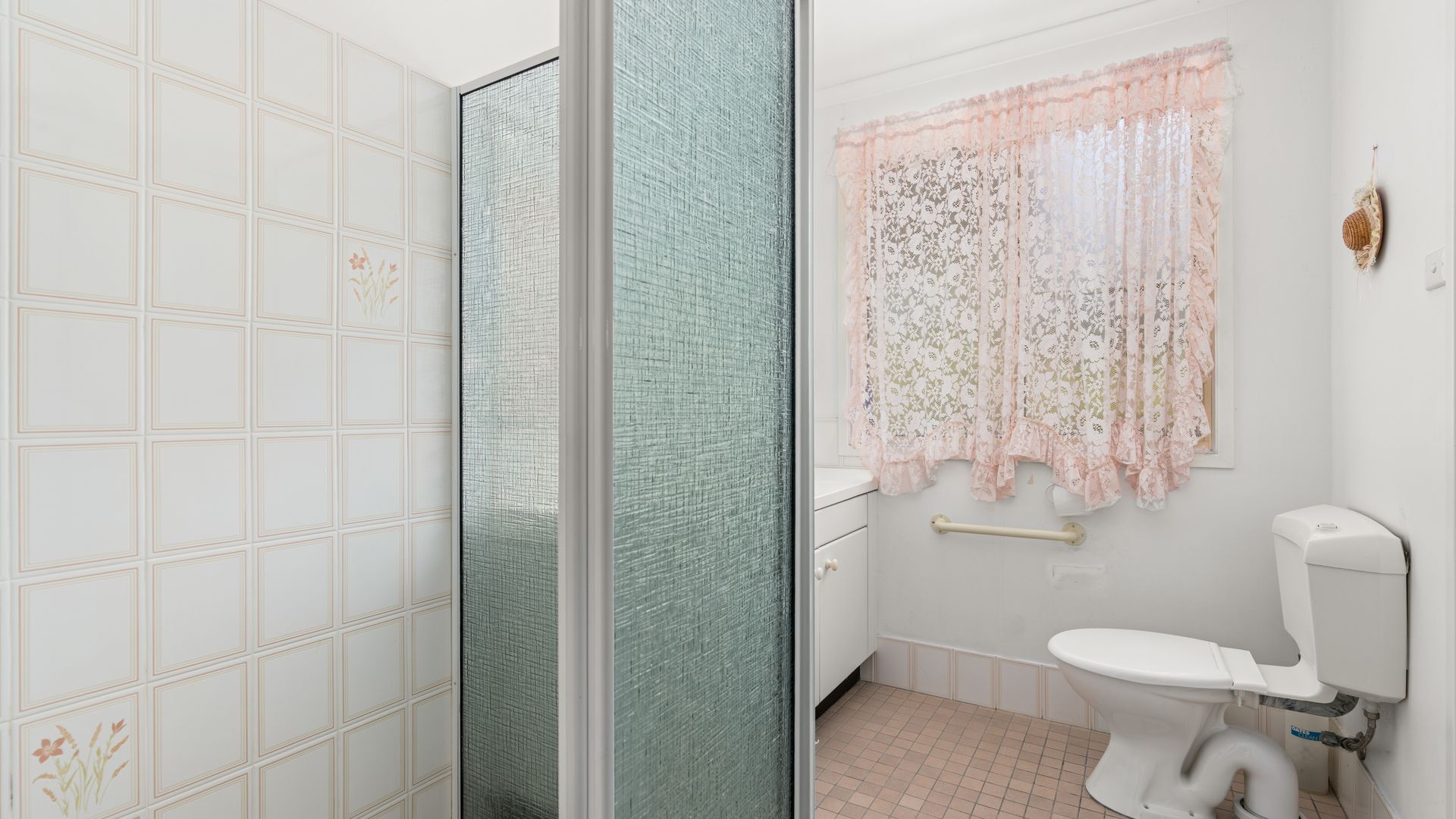 Un baño con inodoro, ducha con puerta de vidrio y ventana con cortinas florales.