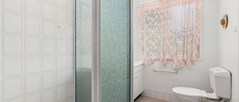 Un baño con inodoro, ducha con puerta de vidrio y ventana con cortinas florales.