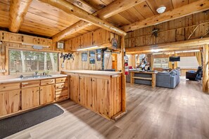 A cabin interior with wooden walls and floors, a kitchen area with a sink and cabinets, a dining area with a table and chairs, and a living room with a sofa and a television. - The Happy Place Cabin (South Fork)