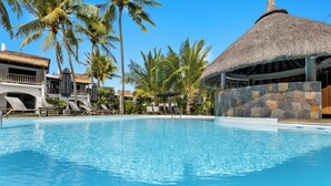 Ein Pool mit Liegestühlen und Sonnenschirmen, umgeben von Palmen und einer Struktur mit Reetdach.