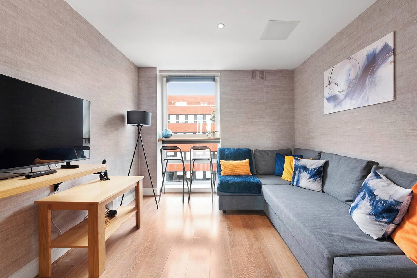 A modern living room with a flat-screen TV, a sofa with colorful cushions, a standing lamp, and a large window offering a view of a brick building.