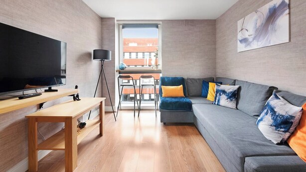 Sala de estar moderna com televisão de tela plana, sofá com almofadas coloridas, luminária de chão e uma grande janela com vista para um edifício de tijolos.