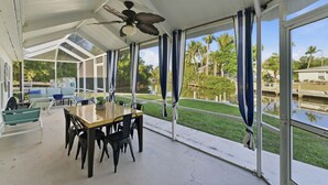 Un espace extérieur abrité, aménagé pour les repas, avec une table et des chaises, de grandes fenêtres et une vue sur un plan d’eau et la végétation.