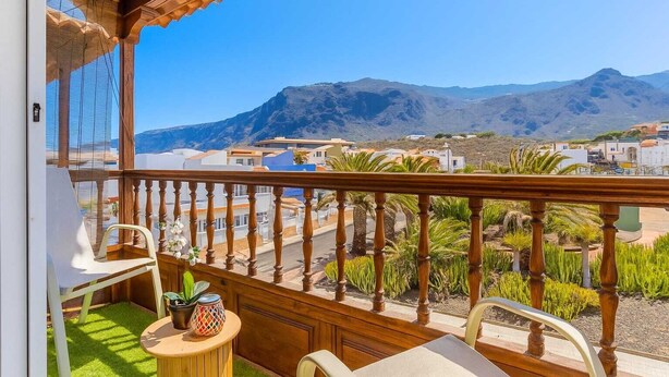 A balcony with a wooden railing, a small table with chairs, and a view of a coastal town with mountains in the background. - TenoHomes Elegant Hideaway in Nature (Puertito de los Silos)