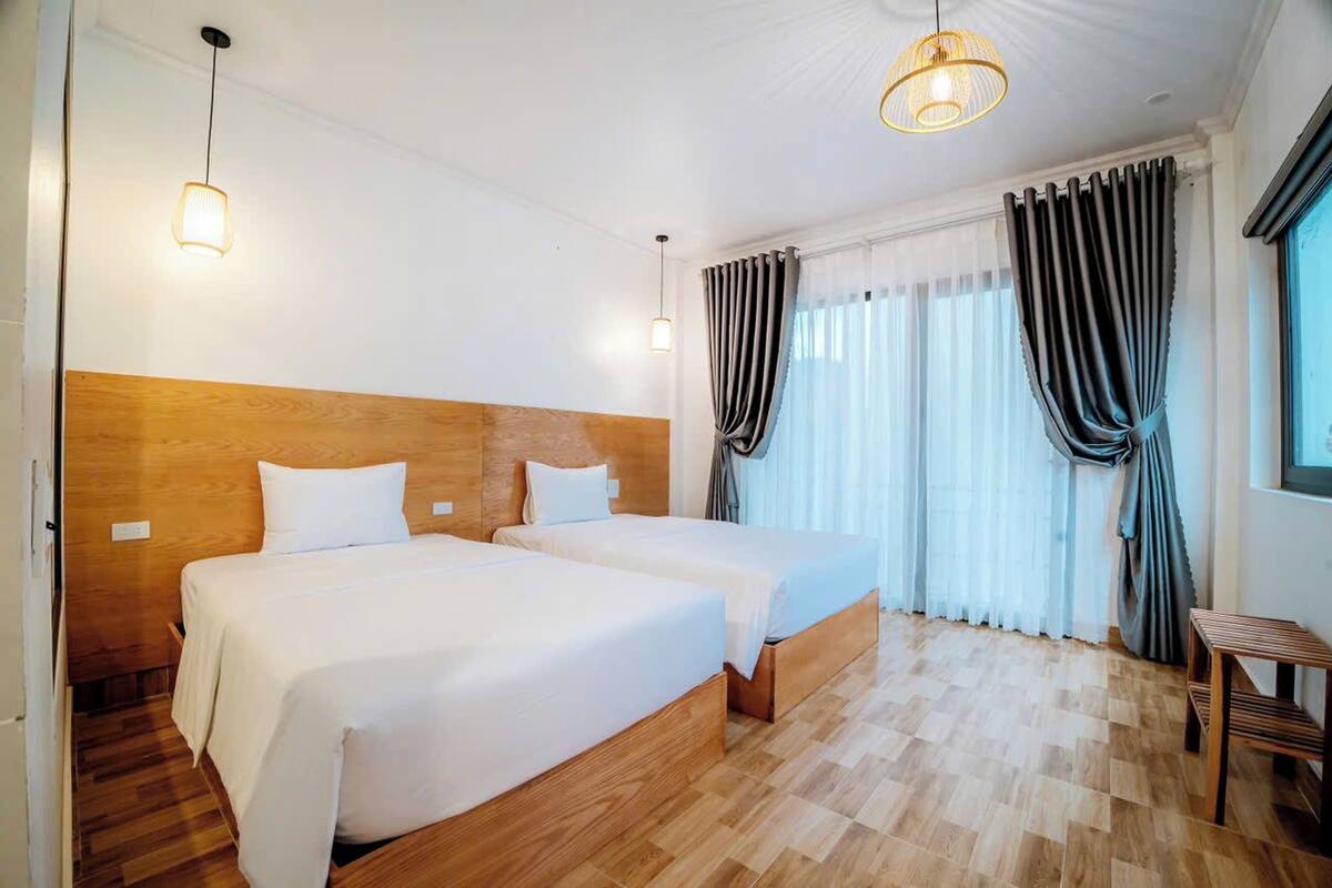 Habitación de hotel con dos camas, cabecero de madera, ropa de cama blanca, una mesita de madera y una ventana con cortinas.