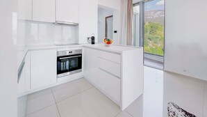 Une cuisine moderne avec des meubles blancs, un four encastré et une grande fenêtre offrant une vue sur la verdure et les montagnes.