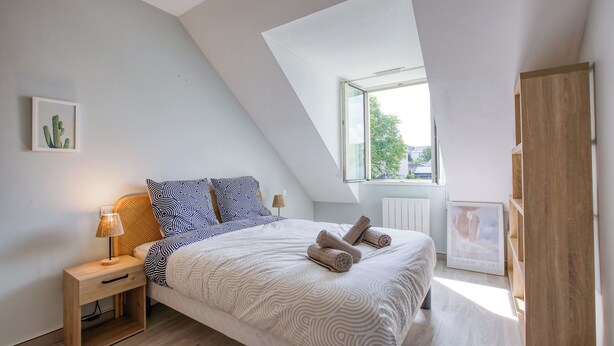 Une chambre avec un plafond en pente, un lit recouvert d’une couvre-lit à motifs, deux oreillers, une table de chevet avec une lampe et un tableau encadré accroché au mur.