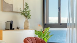 A modern hotel room with a large window offering a view of the ocean, a bedside table with a coffee maker, a plant, and a red chair. - Garajau Sea Side by Atlantic Holiday (Santa Cruz)