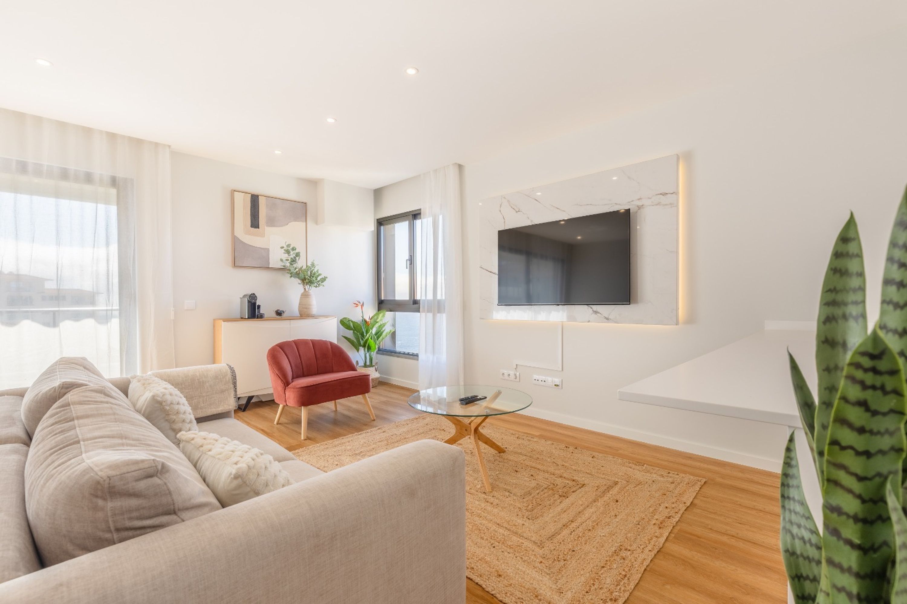 Una sala de estar moderna con un sofá, una butaca roja, una mesa de centro de vidrio y un televisor montado en la pared.