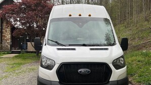 A white Ford van parked on a gravel path with a stone building and trees in the background.