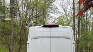 The rear view of a white van parked on a gravel path surrounded by trees.