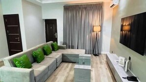 A modern living room with a sectional sofa, a coffee table, a floor lamp, and a flat-screen TV mounted on the wall. - Nook and Cranny Apartment (Abuja)
