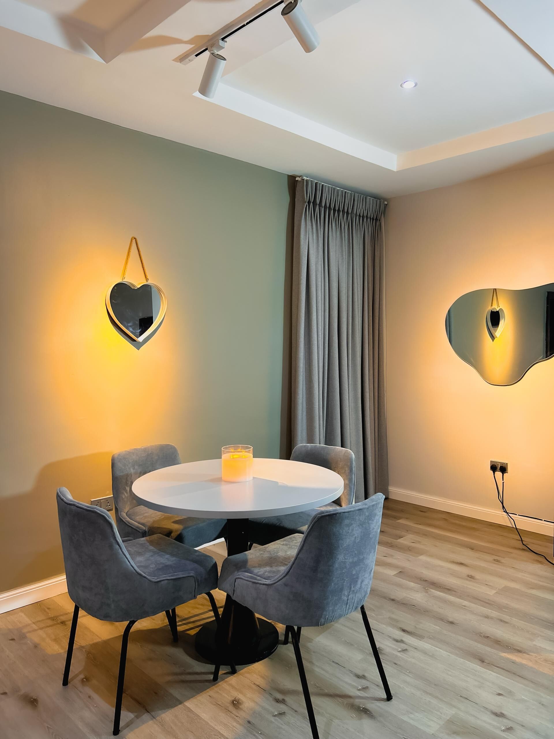 A modern dining area with a round table, four grey chairs, a candle, and a heart-shaped mirror on the wall.