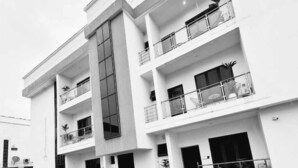 A modern multi-story building with a central glass facade, multiple balconies, and a paved courtyard. - Nook and Cranny Apartment (Abuja)