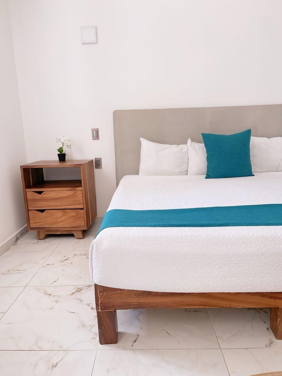 A neatly made bed with a white and teal bedspread, a wooden nightstand with a plant, and a simple wall-mounted light fixture.