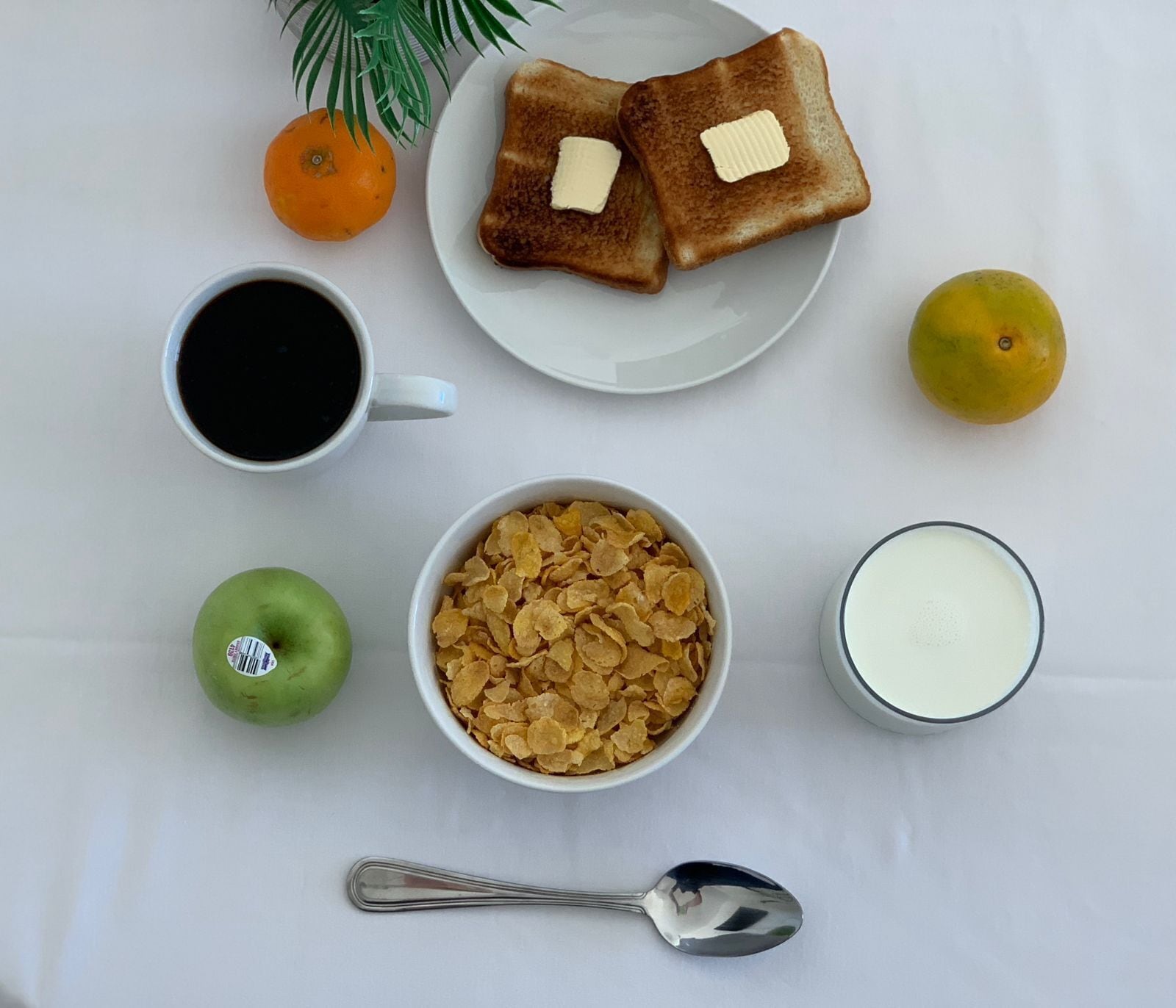En frukostuppsättning med rostat bröd, smör, apelsin, citron, äpple, flingor, mjölk, kaffe och en sked på ett vitt bord.
