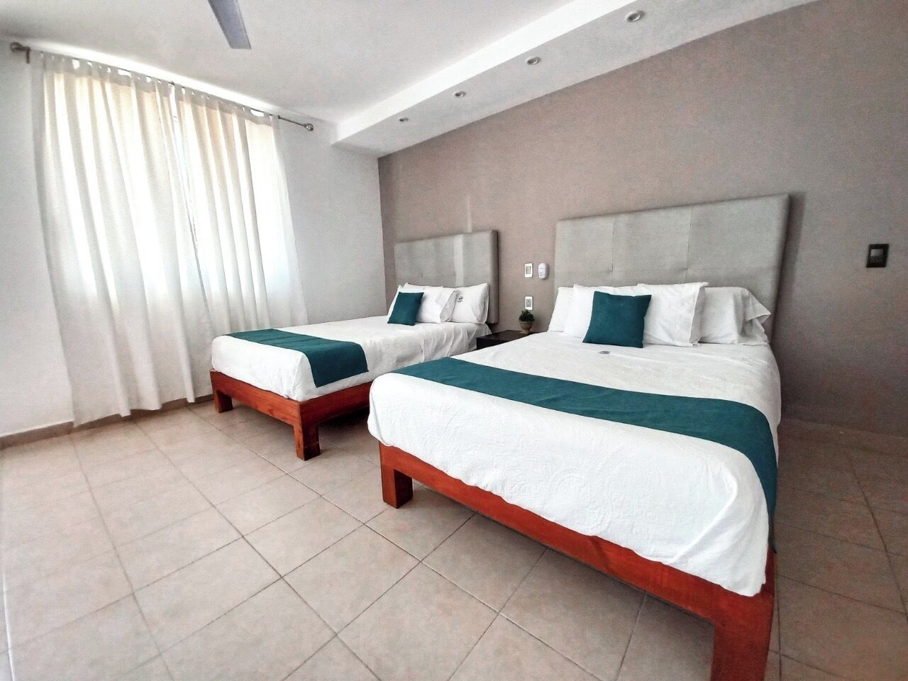 Habitación de hotel con dos camas, un ventanal grande con cortinas y suelo de baldosas.