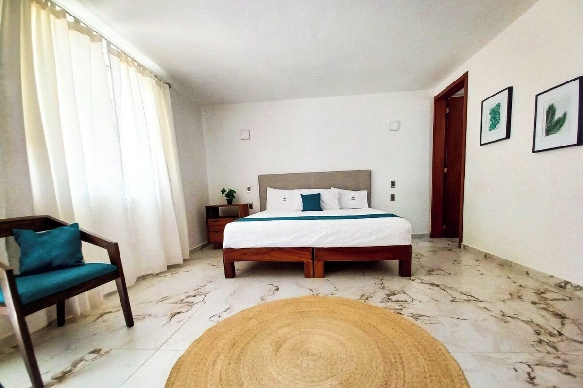 Un dormitorio con cama, una silla, una mesita de noche y una pared decorada con marcos.