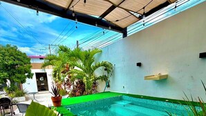 Una piscina con borde de césped artificial verde, rodeada de plantas en macetas, bajo una zona sombreada con estructura de techo metálico.