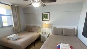 A hotel room with two beds, a ceiling fan, a nightstand, a lamp, and a window with curtains. - Sunrise Studio – Balcony with Ocean Views (Daytona Beach Shores)