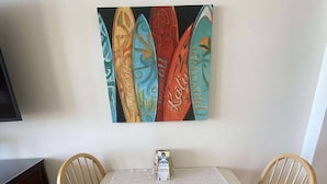 A dining area with a white table, two wooden chairs, and a colorful surfboard painting on the wall. - Sunrise Studio – Balcony with Ocean Views (Daytona Beach Shores)