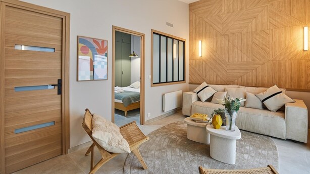 Un salón moderno con sofá, mesa de centro y una silla. Se observa una puerta de madera y una pared con un diseño geométrico.