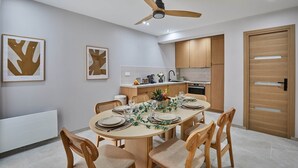Un comedor moderno con una mesa redonda dispuesta para cuatro personas, rodeada de sillas de madera. Un ventilador de techo se encuentra sobre la mesa, y la habitación cuenta con una cocina de armarios de madera y encimera blanca.