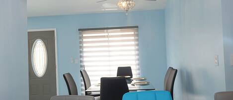 Comedor con una mesa lista para cuatro personas, ventilador de techo y ventana con persianas.