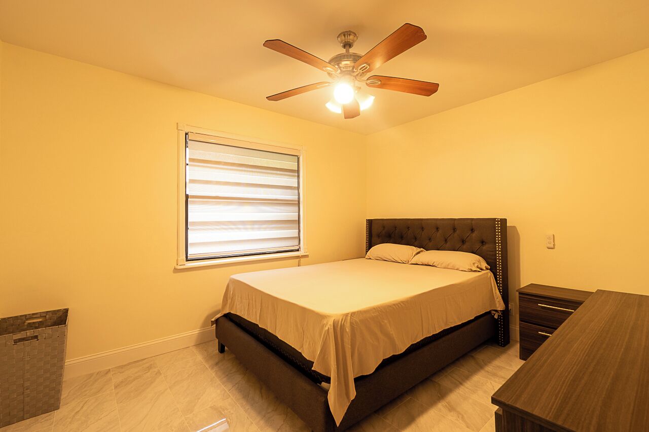 Ein Schlafzimmer mit Bett, Deckenventilator, Fenster mit Jalousien, Nachttisch und Kommode.