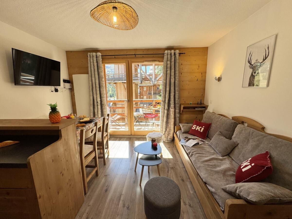 A cozy living room with a sofa, a small table, and a television. There is a wooden dining table and chairs, and a window with curtains.