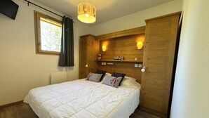 Une chambre avec un grand lit, une tête de lit en bois, deux lampes fixées au mur, une fenêtre avec des rideaux et un téléviseur accroché au mur.