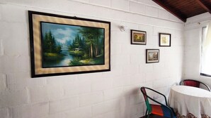 Una habitación con pared de ladrillo blanco, un cuadro con paisaje enmarco, una mesita redonda con mantel blanco y una silla colorida.