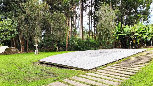 Un área pavimentada con piedras que forman un sendero hacia una plataforma de madera, rodeada de césped y árboles.