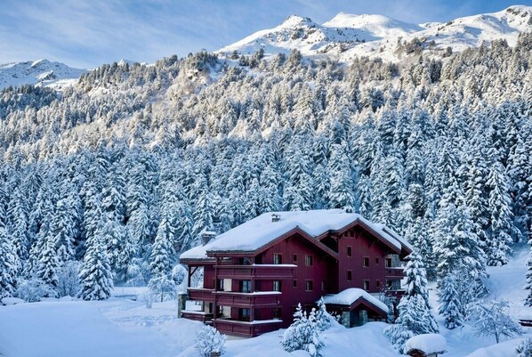 A multi-story chalet with a red facade, surrounded by snow-covered trees and mountains in the background.