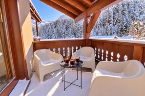 A covered outdoor seating area with two chairs and a table, surrounded by wooden railings, overlooking a snowy landscape with trees.