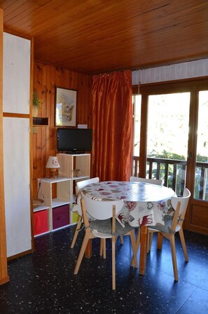 A wooden cabin with a round dining table, chairs, a television, and a balcony with a red curtain.