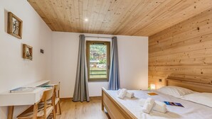 A bedroom with a wooden ceiling, a desk with a chair, a bed with towels, and a window with curtains.