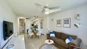 A modern living room with a TV, sofa, and coffee table. There is a dining area with chairs and a table. The room has white walls and a ceiling fan. - Precioso Apartamento Cerca de Marbella con Vistas y Playa (Mijas)