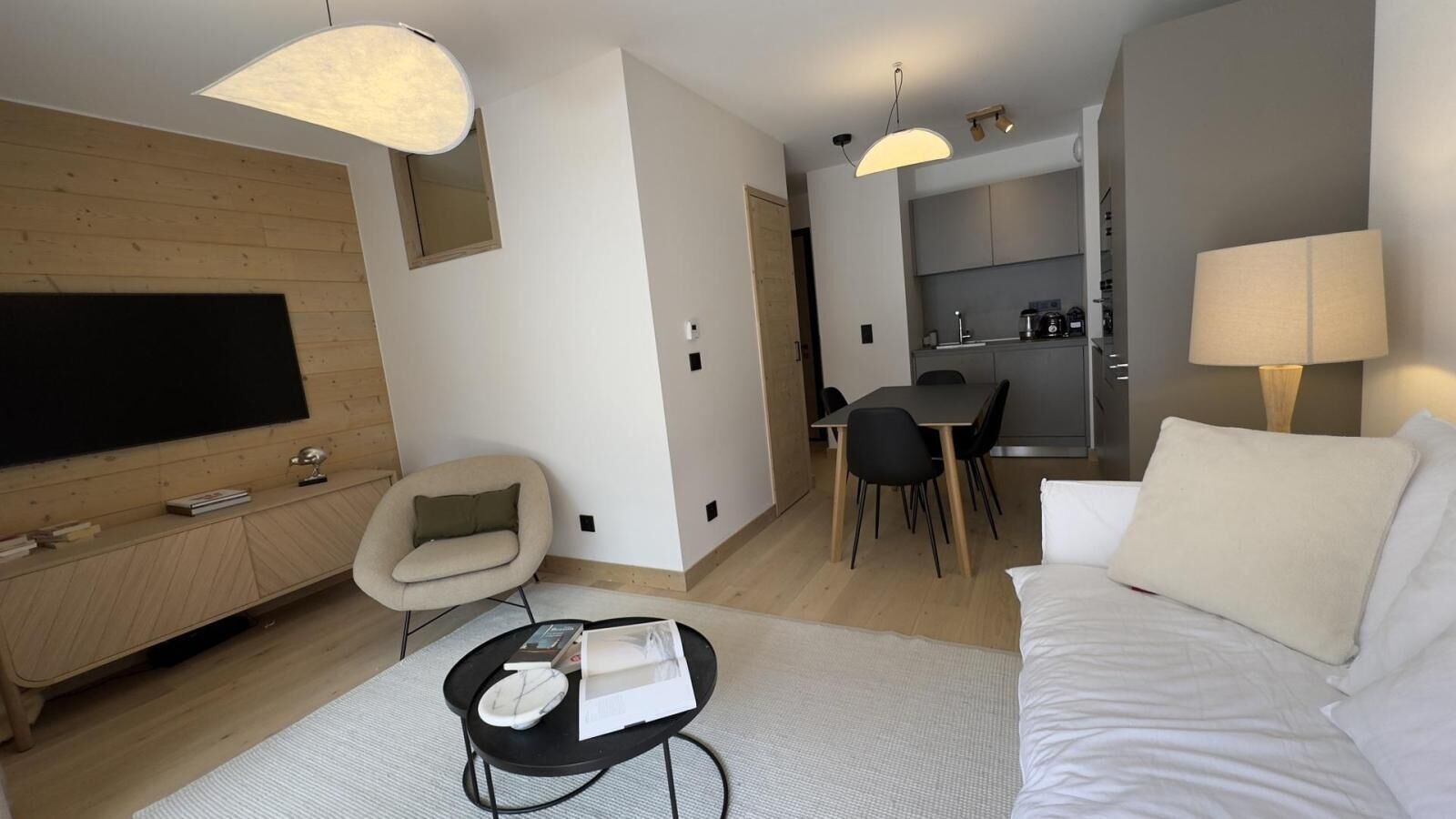 A modern hotel room with a flat-screen TV, a sofa, a dining table with chairs, a kitchen area, and a lamp.