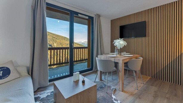 Ett modernt hotellrum med ett matbord i trä, två stolar, en soffa och ett stort fönster med utsikt över ett bergigt landskap.