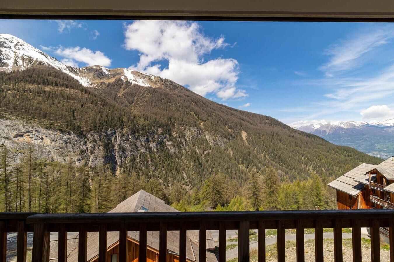 Une vue sur la montagne depuis un chalet en bois doté d’un balcon, entouré de forêts et de sommets enneigés.
